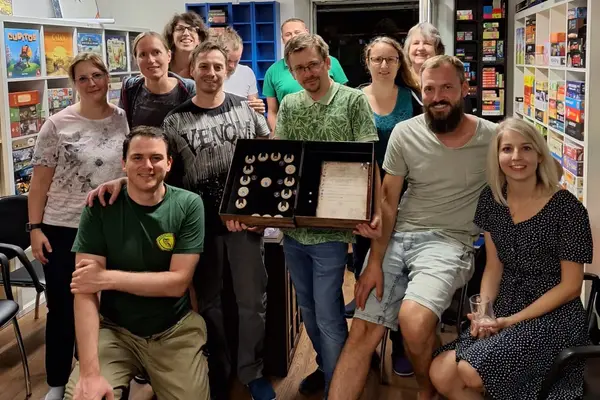 Auf diesem Gruppenfoto werden zwölf Personen gezeigt, die um den Spielleiter des Spiels Blood on the Clocktower versammelt sind und in die Kamera lachen.