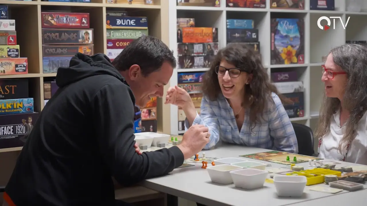 Das Foto zeigt einen Bildausschnitt der QTV Reportage über den Brettspielverein Kufstein, auf welchem ein Mann und zwei Frauen beim Spielen eines Brettspiels gezeigt werden.